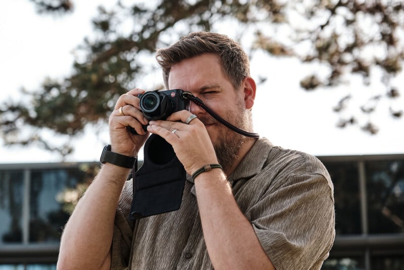 A bearded man wearing a short-sleeved shirt is standing outside, holding a camera up to his face and taking a photo. Trees and a glass building are visible in the background.