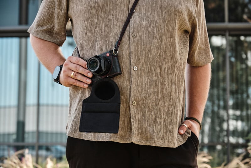 A person wearing a textured brown shirt holds a black Leica camera with a red-stitched strap. The camera hangs in a black protective case.