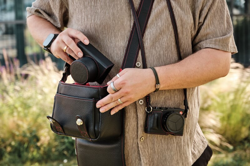 A person wearing a beige shirt and several rings holds a black leather camera bag, placing a lens case inside. Another camera hangs from their shoulder. The background is blurred greenery.
