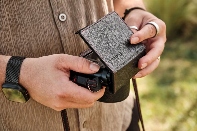 A person wearing a brown shirt and a black watch holds an open leather camera case with a camera partially visible inside. The scene is outdoors with greenery in the background.