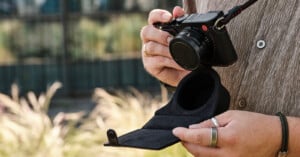 A person wearing rings and a brown shirt holds a black camera in one hand and an open black camera case in the other, standing outdoors with blurred plants in the background.