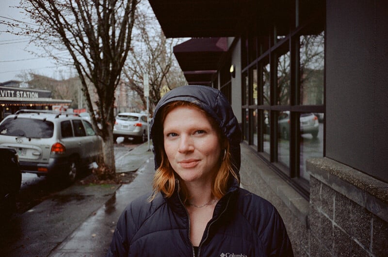 A person with red hair wearing a dark hooded jacket stands on a wet city sidewalk near parked cars and leafless trees on an overcast day.