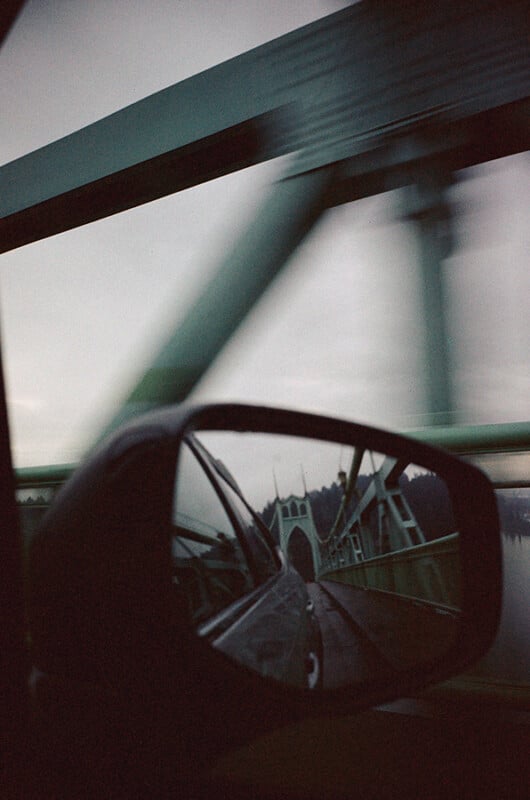 A blurred view of a green steel bridge seen from inside a car, with the bridge reflected clearly in the car’s side mirror under a cloudy sky.