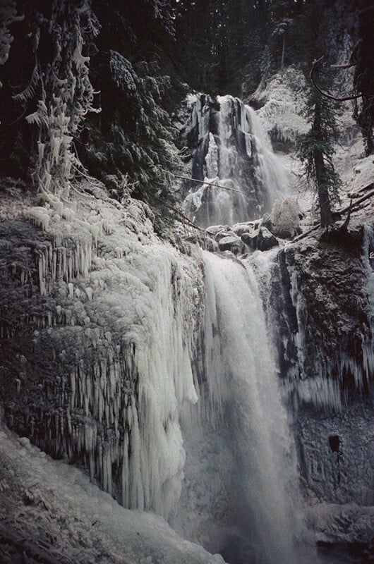 A waterfall flows through a snow-covered forest, with icicles hanging from rocks and trees. The scene is cold and wintry, with frozen surfaces and mist rising from the cascading water.