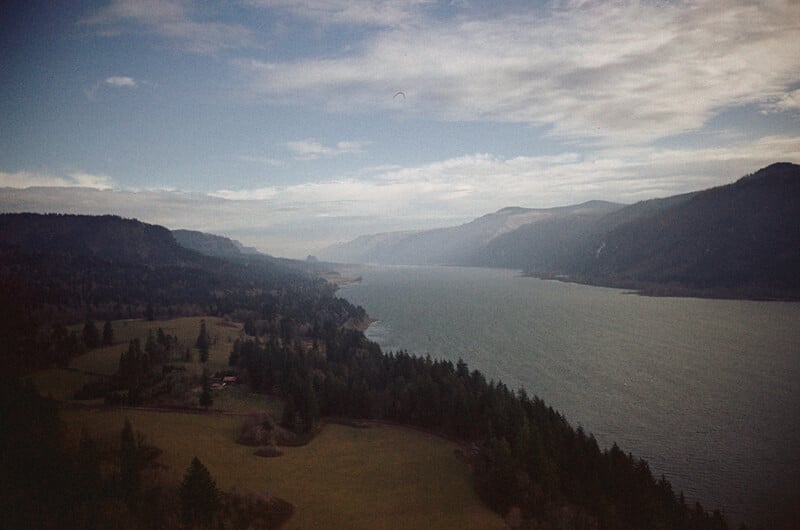 A wide river winds through a verdant landscape of forests and fields, bordered by rolling hills and distant mountains under a cloudy sky.