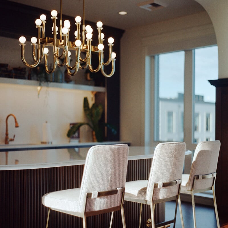 A modern kitchen interior with a white island, three upholstered bar stools, a gold chandelier overhead, a copper faucet, and large windows letting in natural light.