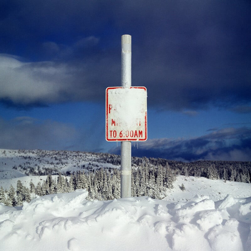 A metal sign partially covered with snow stands in a snow-filled landscape, with trees and hills in the background under a cloudy sky. Only “TO 6:00AM” is clearly visible on the sign.