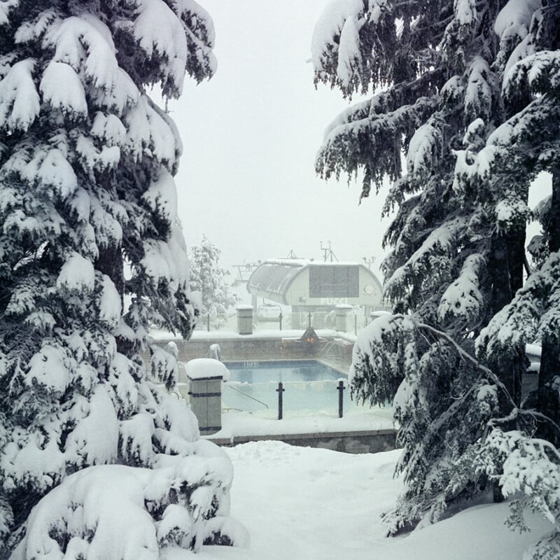 Snow-covered pine trees frame an outdoor pool surrounded by snow, with a ski lift station visible in the background on a foggy winter day.
