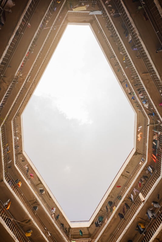 Looking up from the ground floor of a multi-story building with a polygon-shaped opening, balconies lined with railings, and colorful clothes hanging from the upper floors against a bright sky.