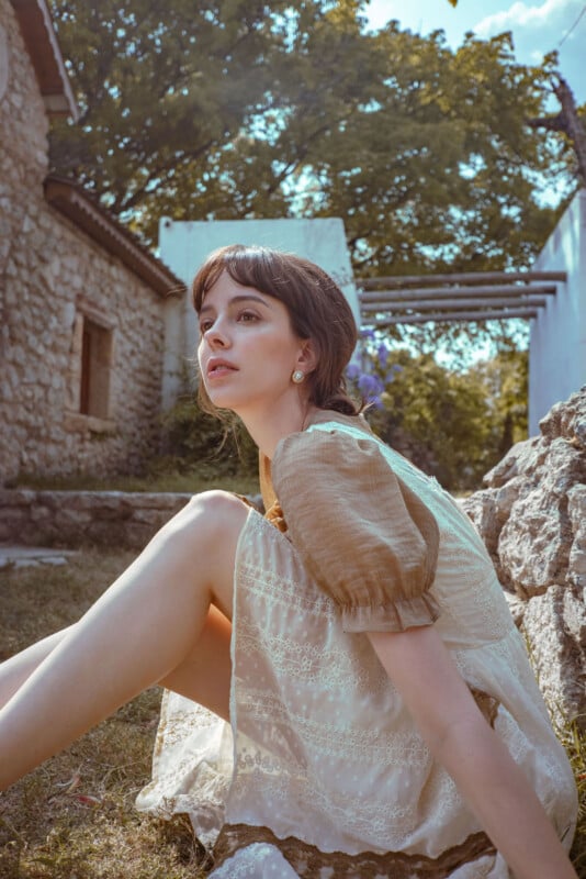 A young woman with brown hair sits outdoors on grass, wearing a light-colored dress with puff sleeves. Stone buildings and green trees surround her, and sunlight filters through the scene. She looks thoughtfully into the distance.