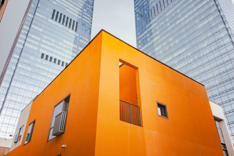 A modern orange building with small windows stands in front of two tall, glass skyscrapers under a cloudy sky, creating a sharp contrast between colors and architectural styles.