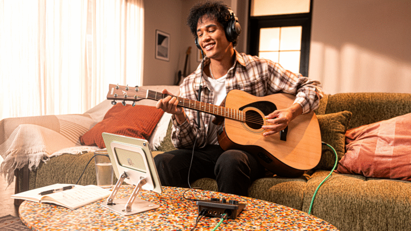 A young person sits on a couch wearing headphones and playing an acoustic guitar. In front of them is a tablet on a stand, a notebook with a pen, and audio equipment on a colorful table.