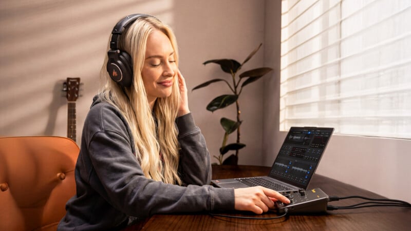 A woman with long blonde hair wearing headphones sits at a desk, smiling with eyes closed, using a laptop and audio mixer. A guitar, plant, and window with blinds are in the background.