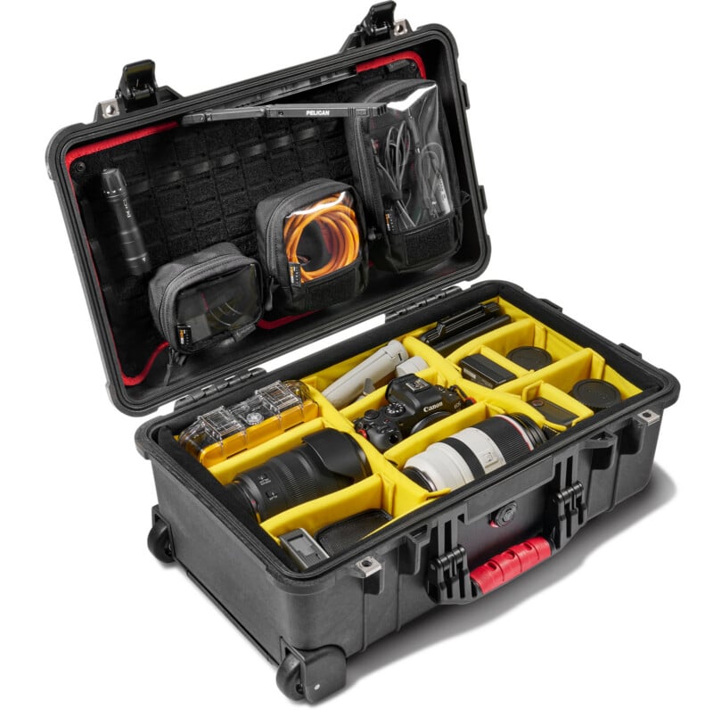 A hard-shell camera case with a yellow padded interior, holding various camera equipment including a DSLR, multiple lenses, batteries, chargers, cables, and accessories. The case lid has zippered mesh pockets for storage.
