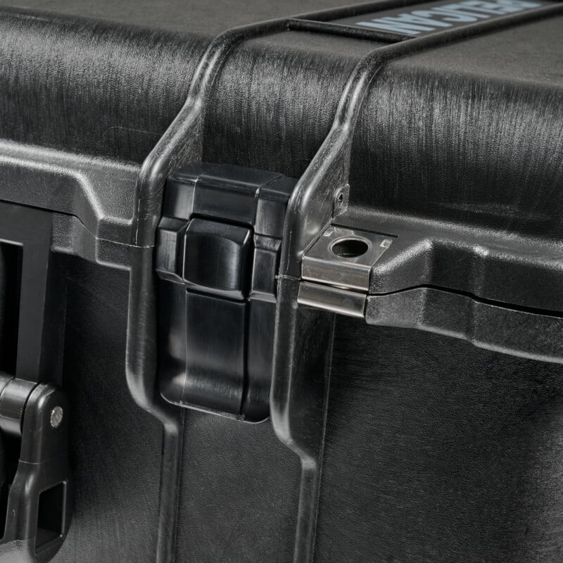Close-up of a black hard plastic case showing a secure latch and metal reinforcement near the hinge, with a textured surface and part of a handle visible at the top edge.