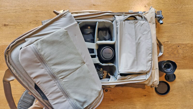 An open beige camera backpack on a wooden surface, showing padded compartments with a camera, lens, and accessories inside. Two lens caps and one detached lens are placed next to the bag.