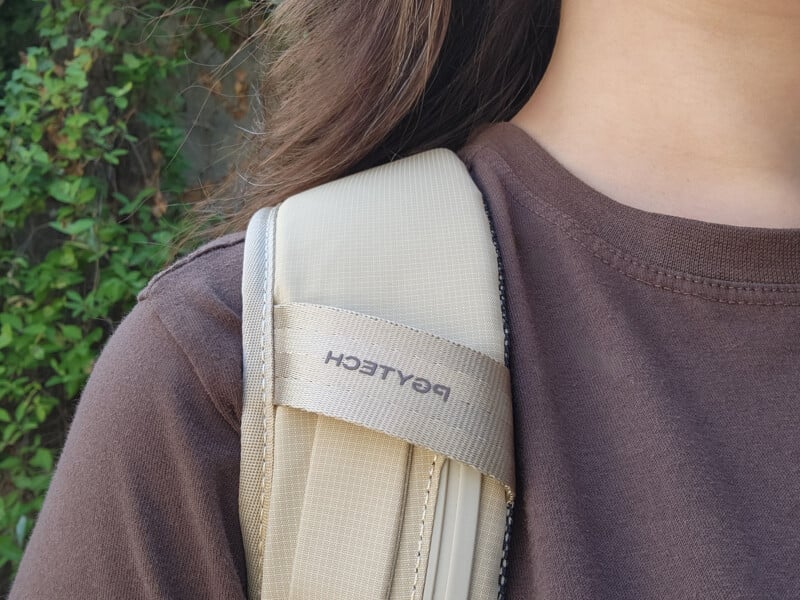 Close-up of a person wearing a brown shirt and a beige backpack with "PGYTECH" written on the strap. Green leaves are visible in the background.