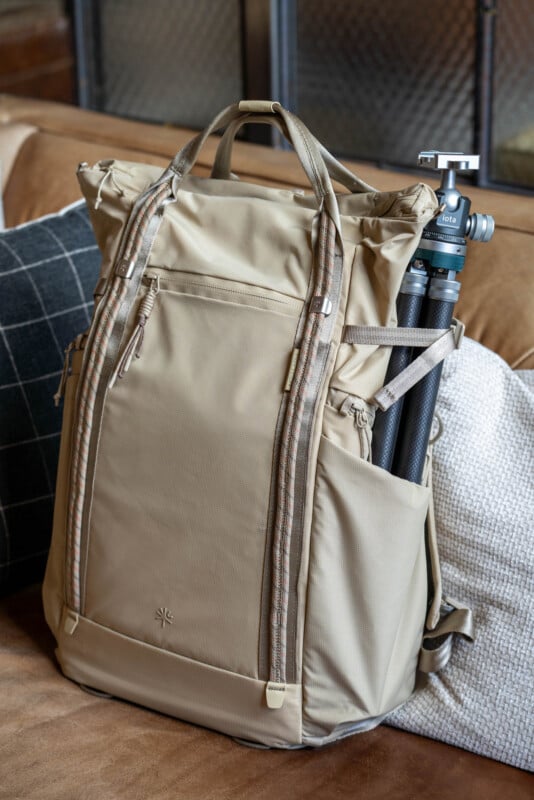 A beige roll-top backpack with patterned straps sits on a couch, holding a black tripod in its side pocket. The backpack is placed between a black-and-white checkered pillow and a plain white pillow.