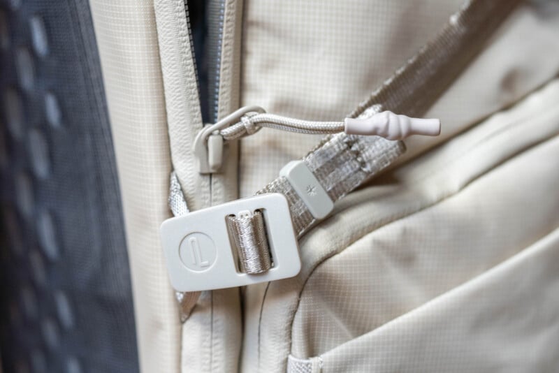 Close-up of a beige backpack showing a zipper with a large plastic pull tab and a thick fabric strap looped through it. The background includes part of a gray mesh panel.