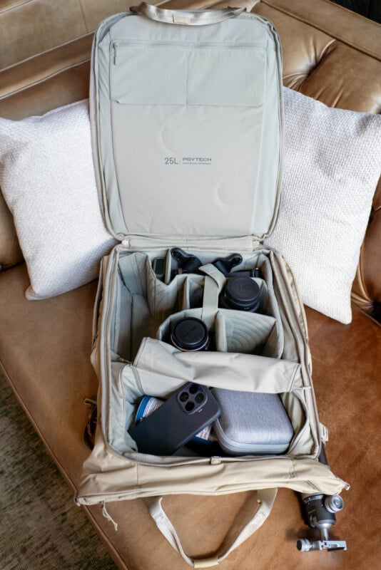 An open beige camera backpack on a tan couch, showing organized compartments with camera lenses, a smartphone, a gray hard case, and a small tripod, along with the label "25L" inside the lid.