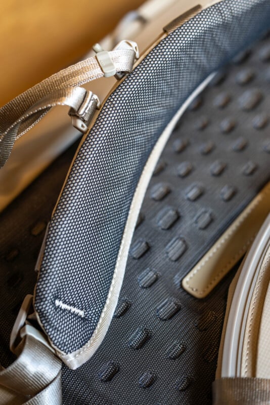Close-up of a textured gray and beige laptop bag or backpack, showing the woven fabric strap, plastic buckle, and patterned surface with raised dots and stitched seams.