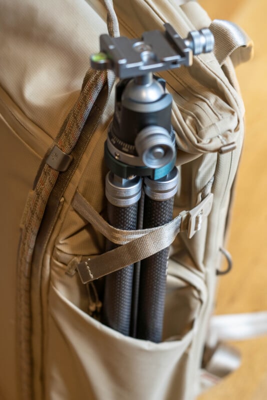 A close-up of a beige backpack with a black tripod secured in a side pocket using a strap. The backpack is resting on a wooden surface.