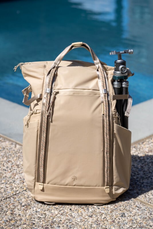 A beige backpack with multiple pockets and two top handles is standing on a poolside patio, with a black tripod stored in one of its side pockets. The pool water is visible in the background.