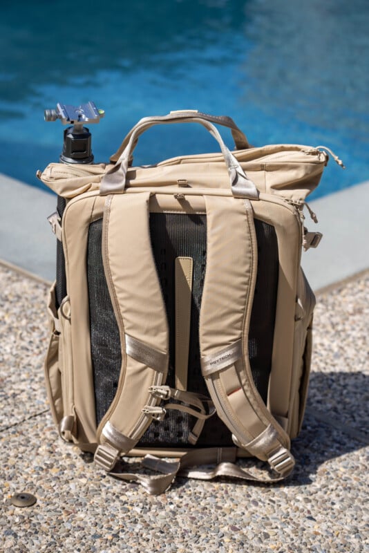 A tan-colored backpack with padded shoulder straps sits on a pebbled surface next to a swimming pool. A camera tripod is secured in a side pocket.