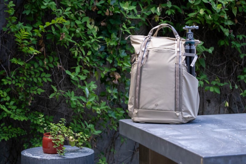 A beige backpack with a tripod in its side pocket sits on a gray table outdoors, near a small round table holding a potted plant. Green vines cover the stone wall in the background.