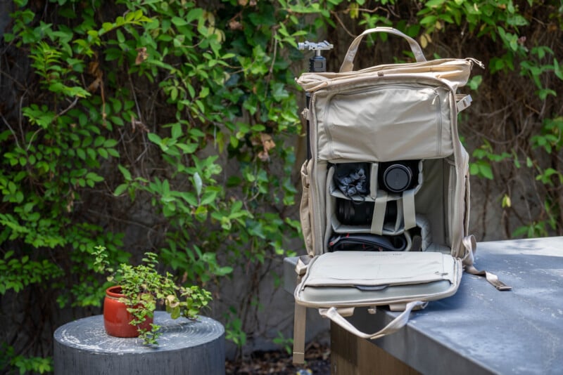A beige camera backpack sits open on a table outdoors, revealing a camera and accessories inside. Nearby, a small planter with green plants sits on a round table, with leafy vines in the background.
