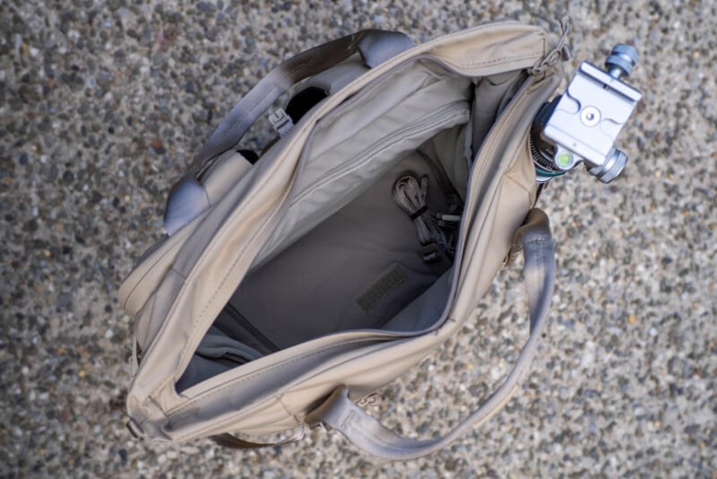 A beige tote bag seen from above, showing its empty main compartment and interior pockets. A metal tripod is attached to the bag’s side. The bag rests on a textured, gravel-like surface.