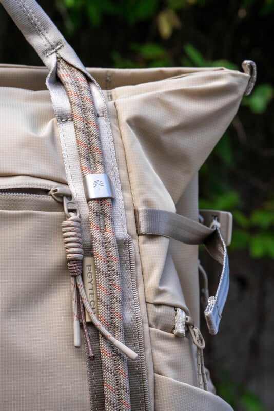Close-up of a beige backpack with textured fabric, visible zippers, and a strap featuring a small metal tag labeled "PGYTECH." Green foliage is blurred in the background.