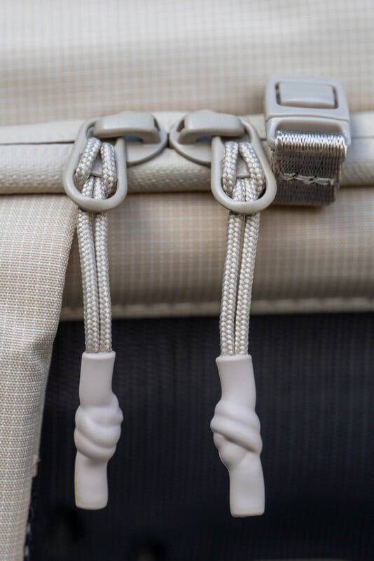 Close-up of two beige zipper pulls with knotted cords and plastic ends on a textured fabric bag, next to a matching strap with a metal buckle.