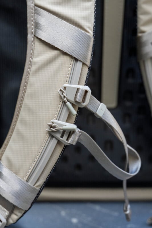Close-up of beige backpack shoulder straps with two plastic adjustment buckles and grey nylon straps, set against a blurred dark background.