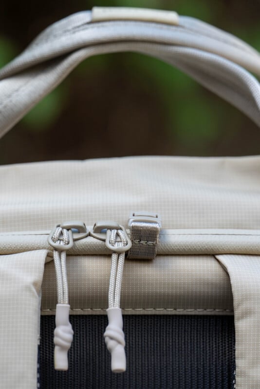 Close-up of the top section of a beige backpack, showing two zippers with white pull cords and a metal clip in the center. The background is blurred and green.