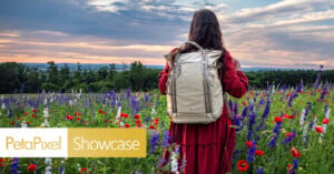 A person in a red dress stands in a colorful wildflower field at sunset, wearing a beige backpack. The sky is partly cloudy, and the PetaPixel Showcase logo appears in the lower left corner.