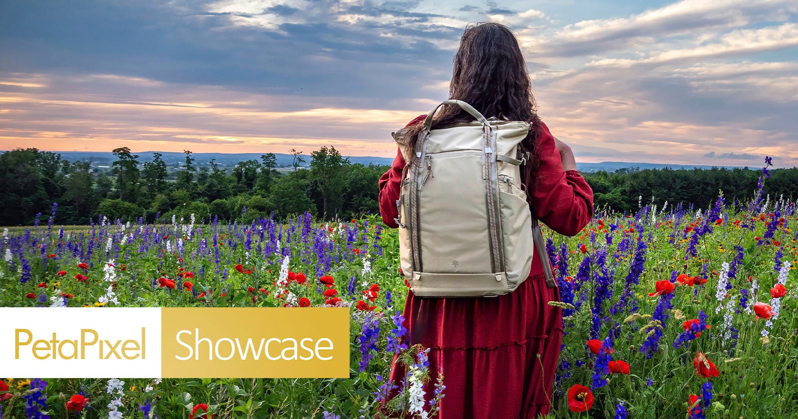 A person in a red dress stands in a colorful wildflower field at sunset, wearing a beige backpack. The sky is partly cloudy, and the PetaPixel Showcase logo appears in the lower left corner.