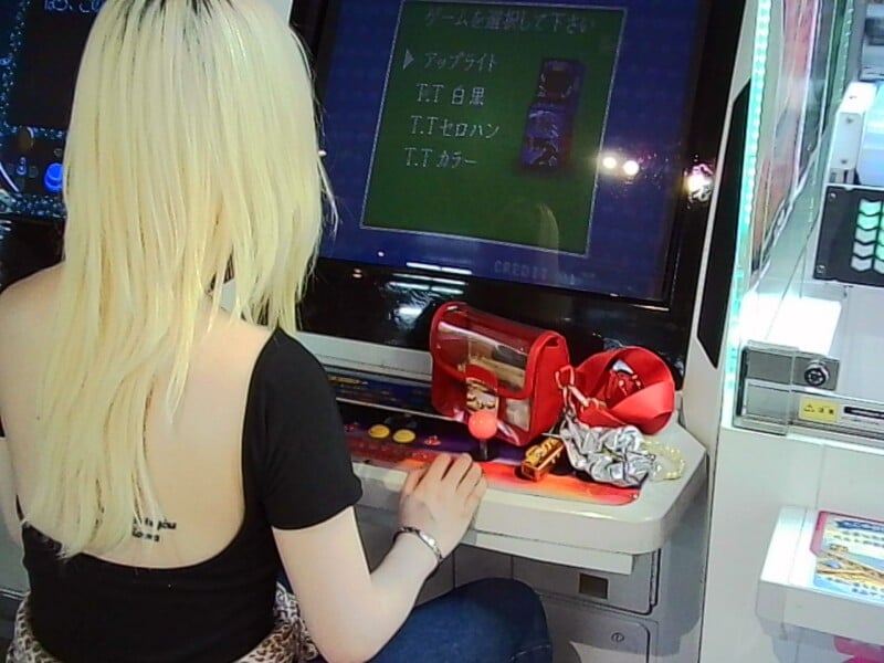 A person with long blonde hair plays an arcade game, facing the screen. There are various items, including a red pouch and crumpled paper, on the arcade machine near the controls.