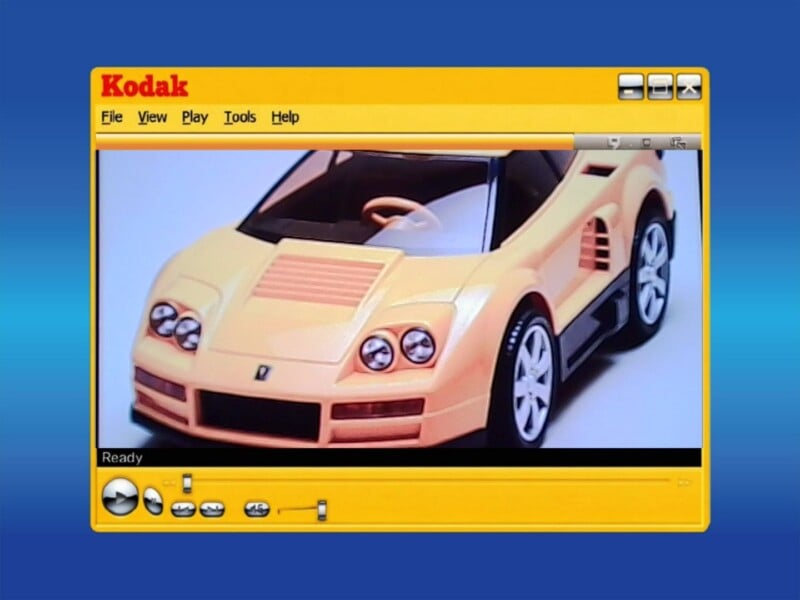 A digital photo of a yellow toy sports car is displayed on a vintage Kodak software interface, set against a blue background. The car has black detailing, white wheels, and is viewed from the front left angle.