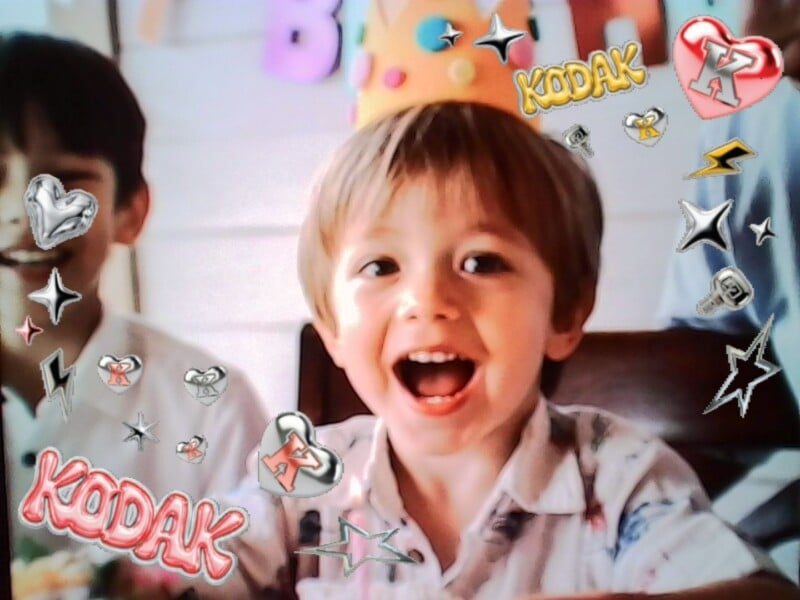 A young child wearing a crown and smiling widely at a birthday party, with colorful "Kodak" stickers and star graphics overlaid on the image. Another child is partly visible in the background.