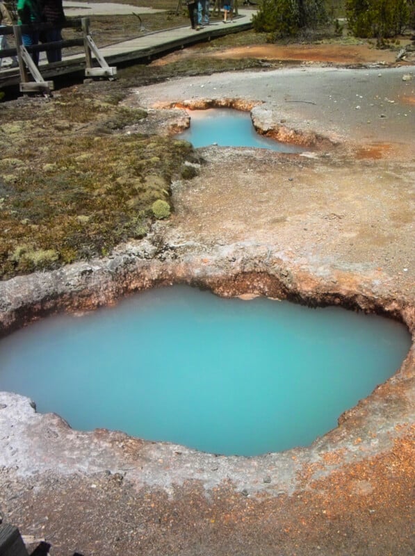 Two bright blue hot spring pools surrounded by rocky ground and sparse vegetation, with a wooden boardwalk and a few people visible in the background.