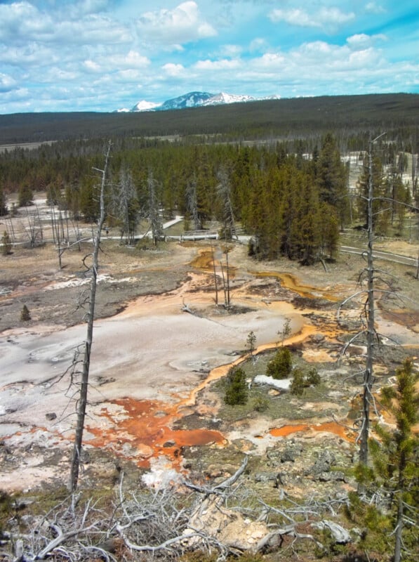 A geothermal area with orange and white mineral deposits, surrounded by sparse trees and forest. Snow-capped mountains are visible in the distance under a partly cloudy sky.