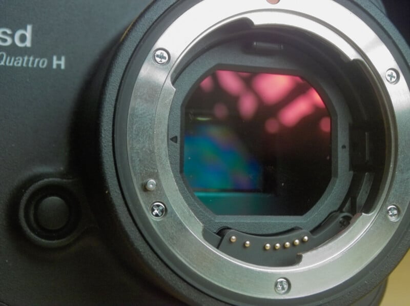 Close-up of a digital camera body showing the lens mount and the exposed image sensor inside, with some colorful reflections visible on the sensor surface.