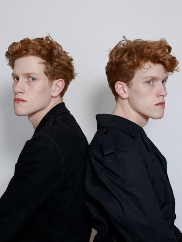Two young men with curly red hair and fair skin sit back to back, both wearing black jackets, against a plain white background. Their expressions are serious and they look in opposite directions.
