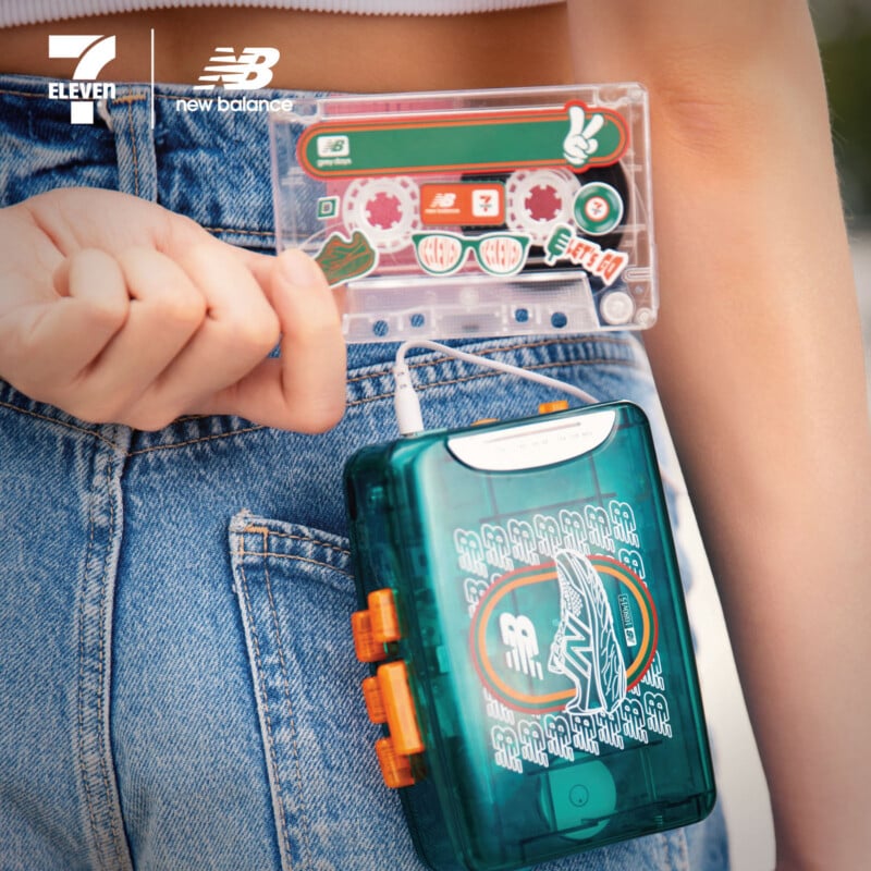 A person in jeans clips a transparent green portable cassette player with New Balance branding to their waistband, holding a matching cassette tape. The 7-Eleven and New Balance logos appear in the upper left corner.