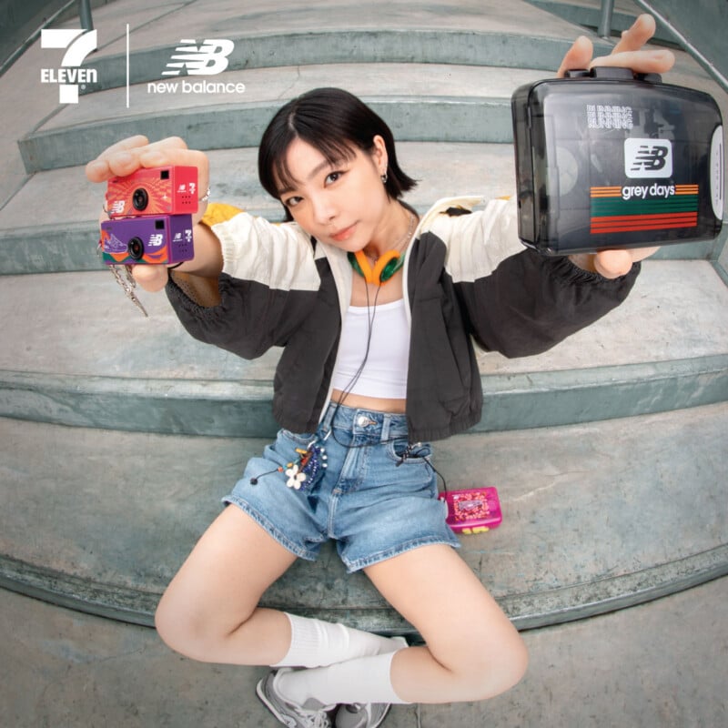 A young woman sits on concrete steps, holding up vintage cameras toward the viewer. She wears a black and white jacket, denim shorts, white socks, sneakers, and headphones. 7-Eleven and New Balance logos are visible in the corner.