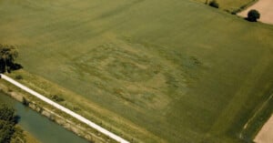 Aerial view of a large green field with a faint, lighter rectangular patch of different shades in the grass, possibly forming a subtle pattern or design. A road and a canal run along the edge of the field.