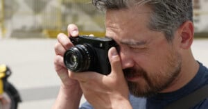 A man with short hair and a beard is holding a black camera up to his face, looking through the viewfinder as he takes a photo outdoors in bright daylight.