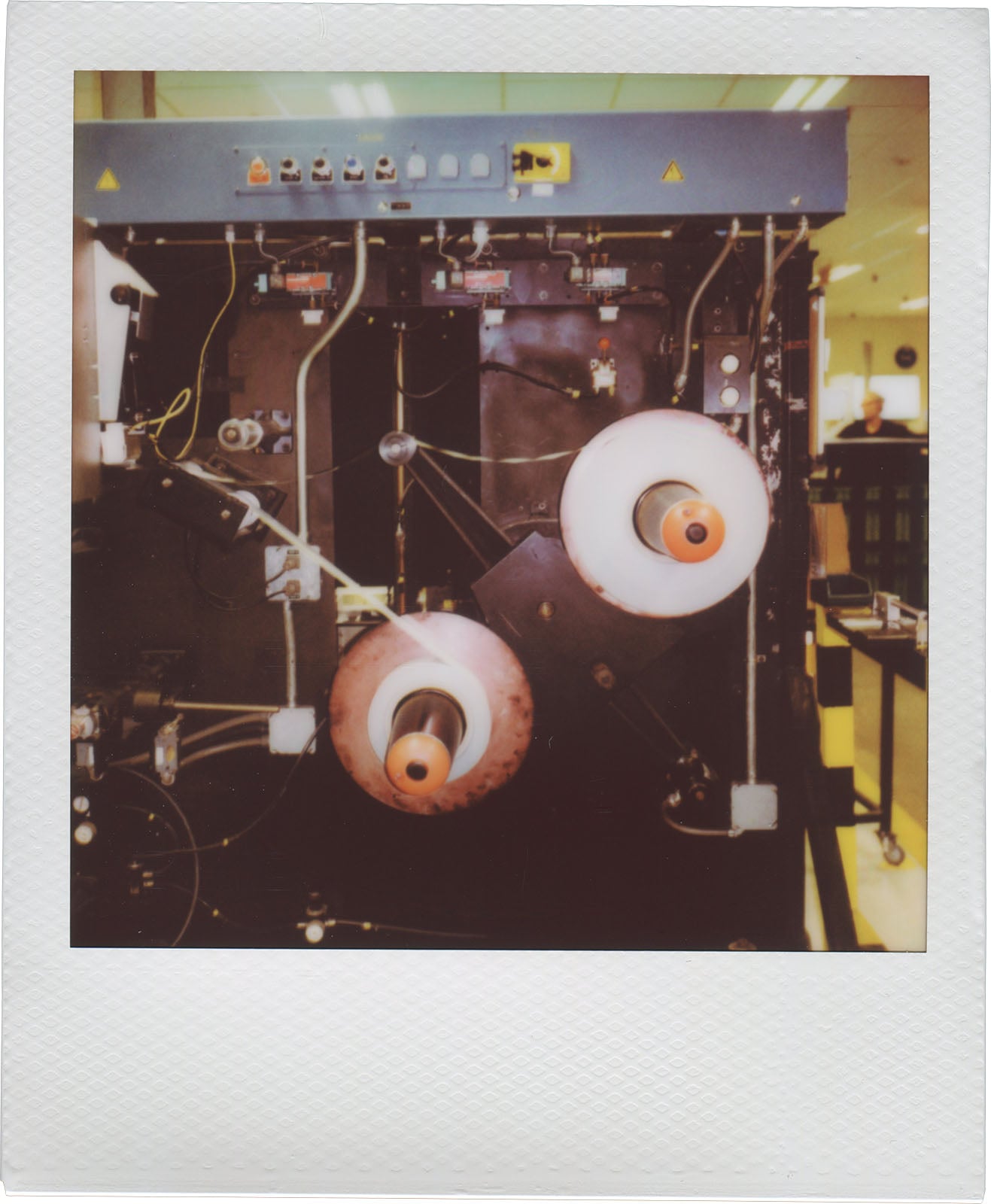 A vintage Polaroid photo showing an industrial machine with exposed gears, wires, and two large spools of material, set in a factory or workshop environment.
