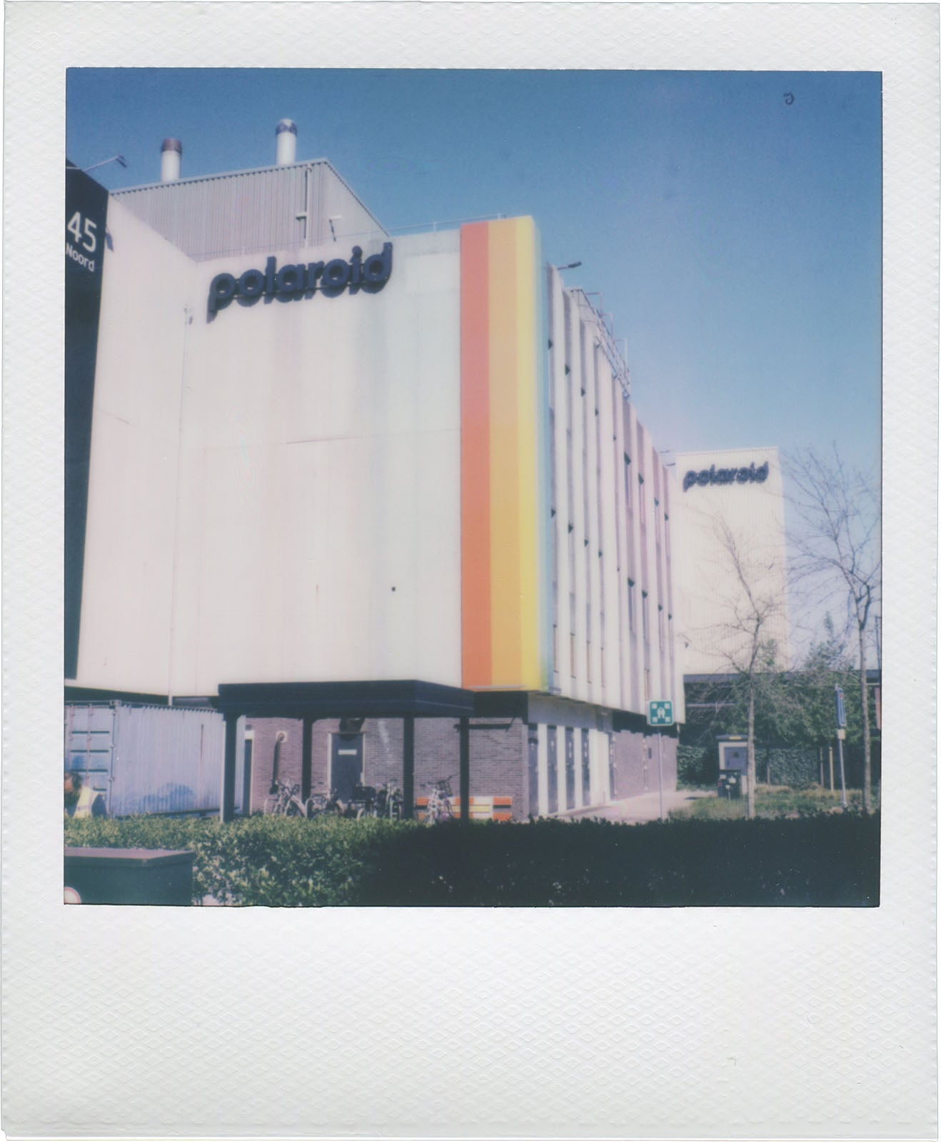 A white building with "polaroid" signage and vertical stripes in yellow, orange, and red stands on a sunny day. Trees, shrubs, and a bicycle are visible in front of the building.